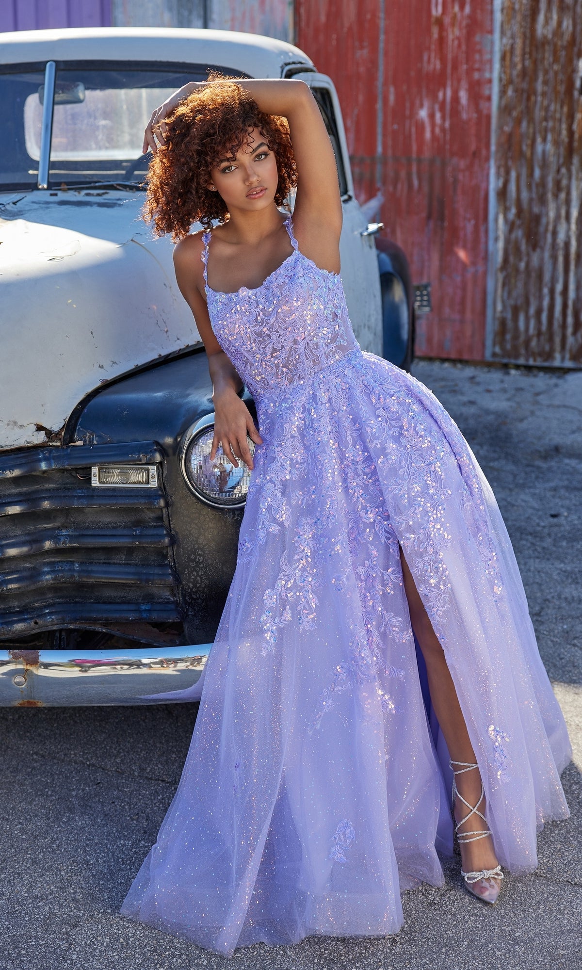 Woman in a Glitter-Tulle Ellie Wilde Long Prom Dress EW35114 with a high slit poses by a weathered vintage car in an outdoor, industrial setting.